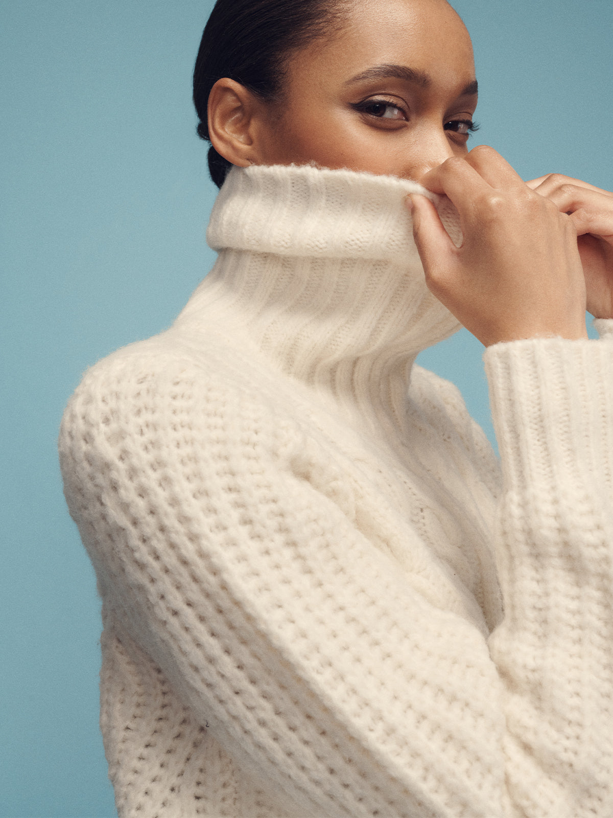 A person with dark hair is partially covering their face with the high collar of a chunky chalk-colored cable zip sweater jacket from Sanctuary Clothing. They are looking sideways against a light blue background.