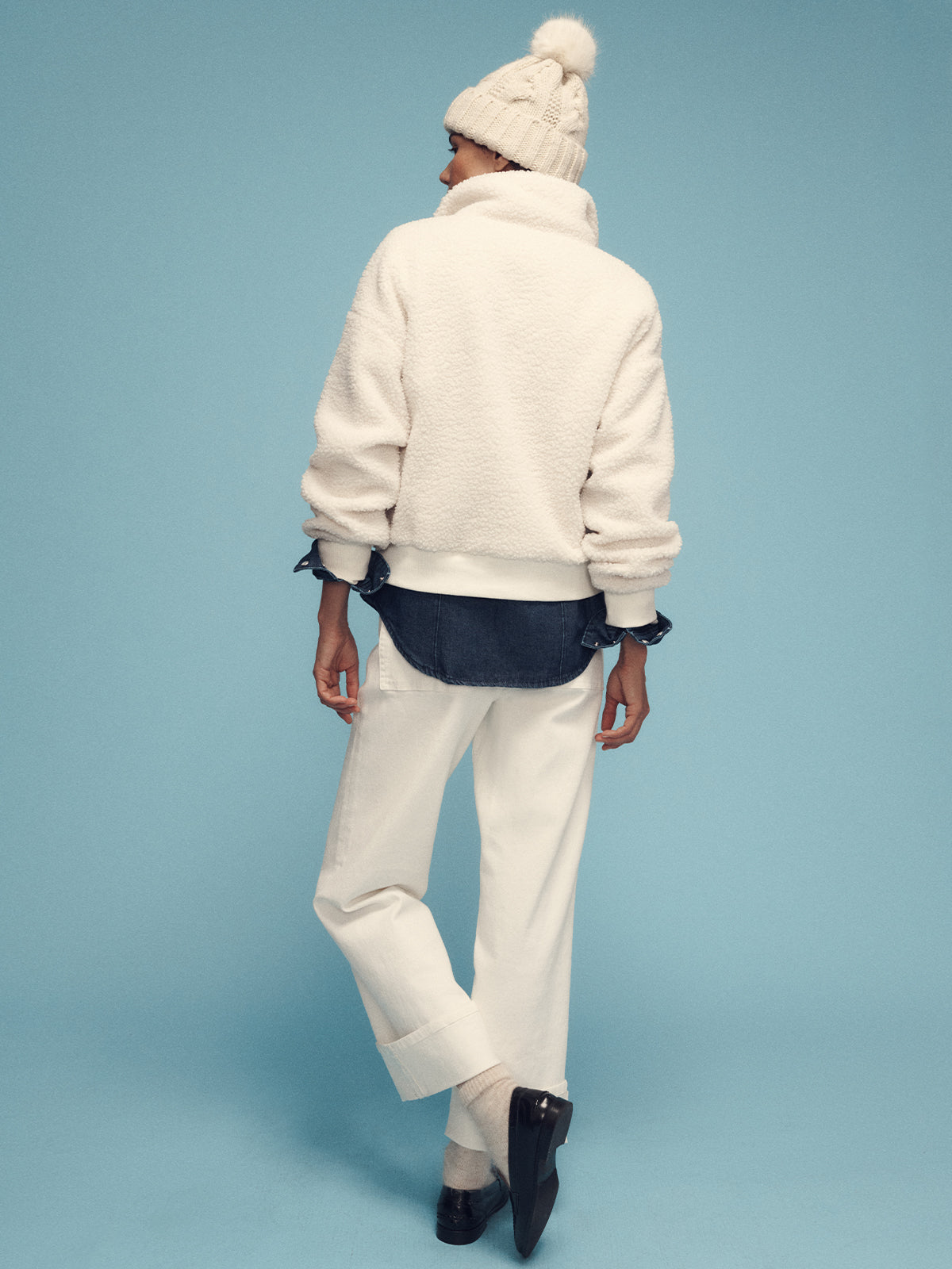 A person wearing the Sanctuary Clothing's alpine sherpa half zip popover in chalk, paired with white pants and a knit beanie adorned with a pom-pom, stands against a light blue background. They are facing away and have layered their outfit with a dark shirt beneath the popover.