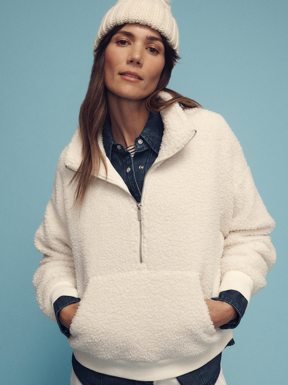 A person wearing the Sanctuary Clothing alpine sherpa half zip popover in chalk and a knitted beanie poses against a blue background. They have their hands in the pockets of the pullover and wear a denim shirt underneath.