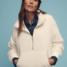 A person wearing the Sanctuary Clothing alpine sherpa half zip popover in chalk and a knitted beanie poses against a blue background. They have their hands in the pockets of the pullover and wear a denim shirt underneath.