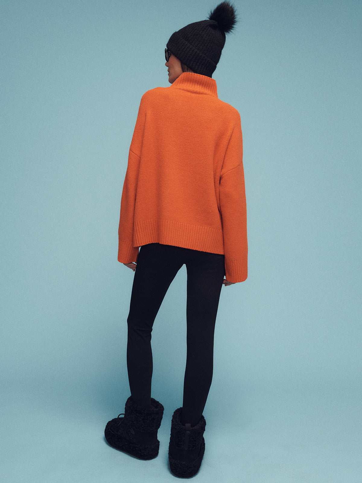 Standing against a blue background, the individual is modeling an "apres intarsia logo sweater ski orange chalk" by Sanctuary Clothing, paired with black leggings, fuzzy black boots, a wooly black beanie adorned with a pom-pom, and sunglasses while facing away from the camera.