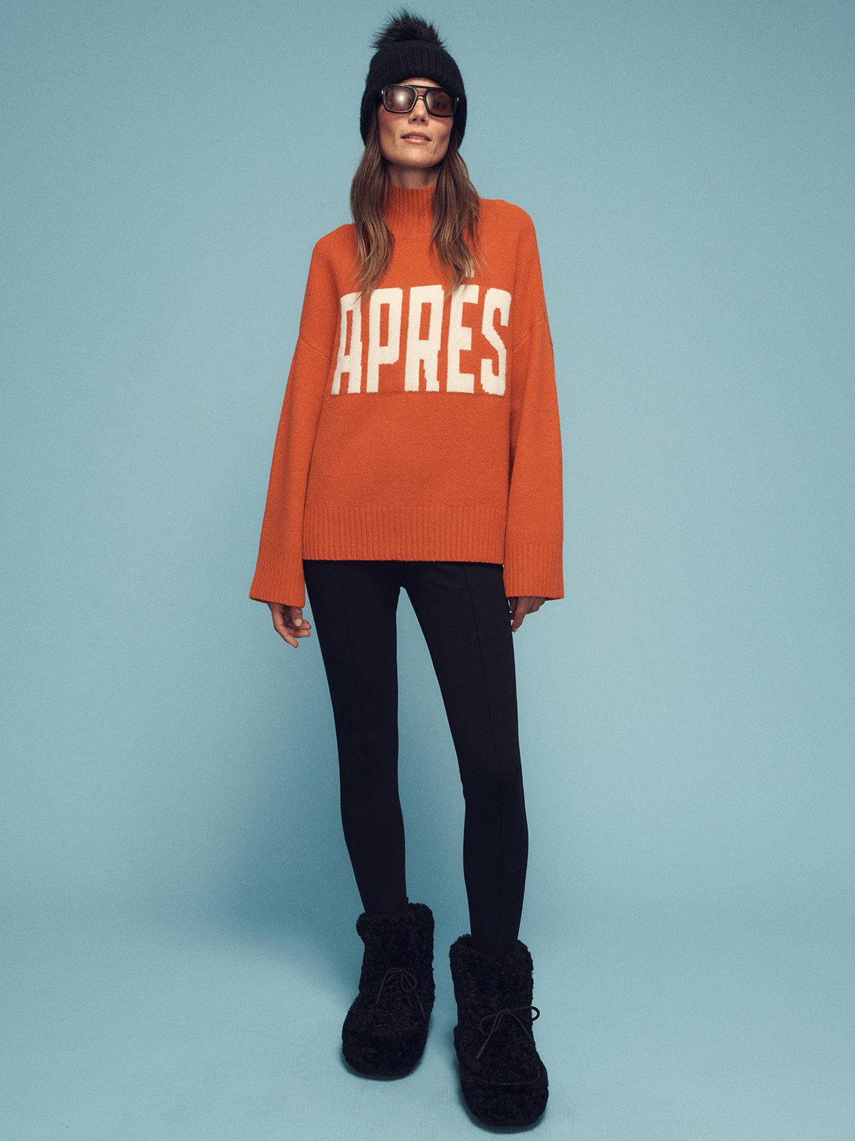 A person wearing an orange sweater with "APRÈS" written on it, designed by Sanctuary Clothing, stands against a light blue backdrop. They are also wearing black leggings, fuzzy black boots, a black hat, and sunglasses.