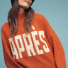 A woman poses against a blue background, wearing the "Apres Intarsia Logo Sweater Ski" in orange chalk by Sanctuary Clothing, paired with a black beanie and dark sunglasses.