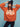 A person dressed in a vibrant orange "apres intarsia logo sweater" by Sanctuary Clothing, featuring the word "APRÈS" in white, paired with black pants, a black knit beanie, and oversized sunglasses, strikes a pose against a blue background with their hands on their hat.