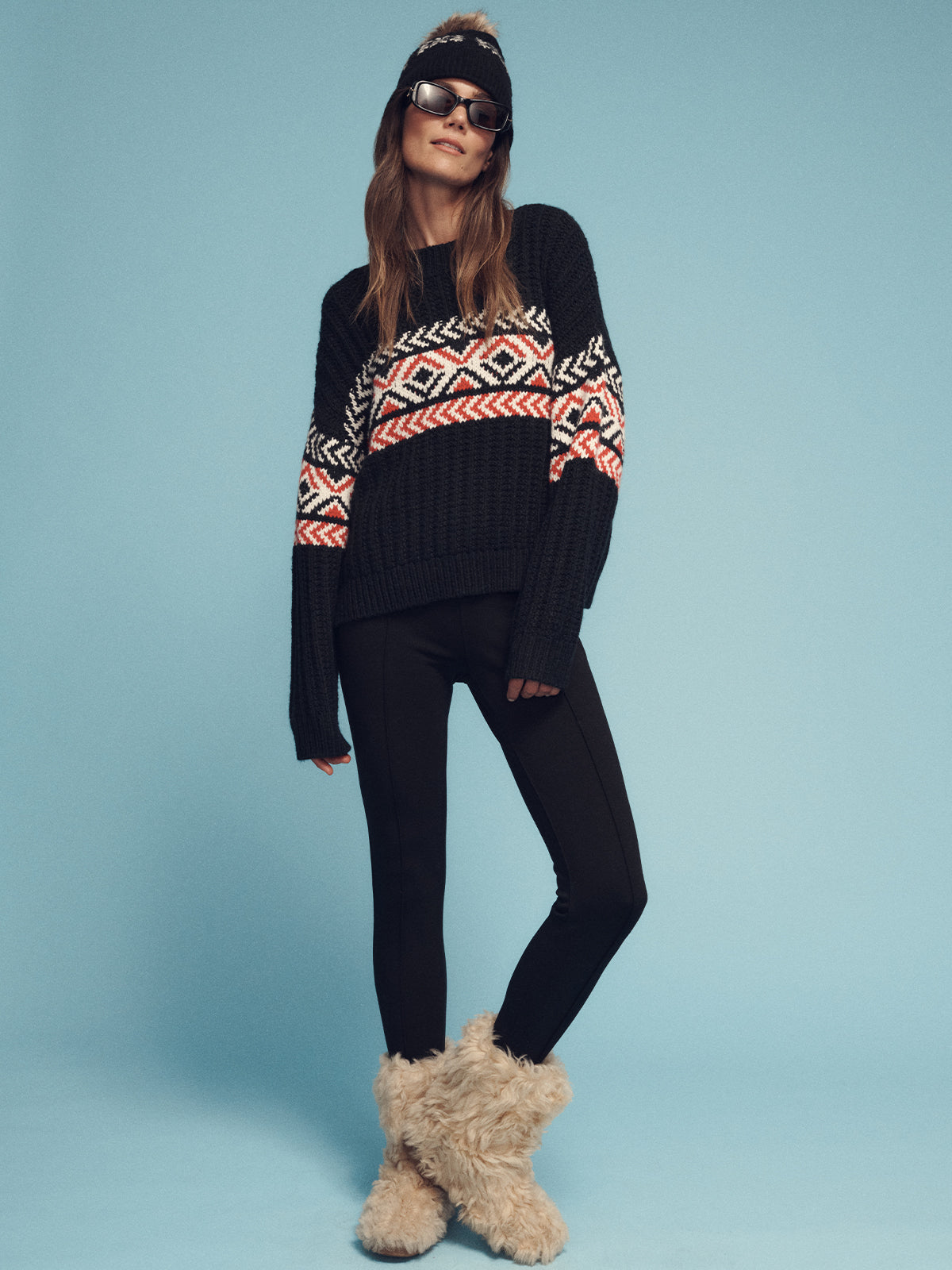 A person wearing the intarsia sweater black multi by Sanctuary Clothing, paired with black leggings, furry beige boots, sunglasses, and a beanie poses confidently against a light blue background.