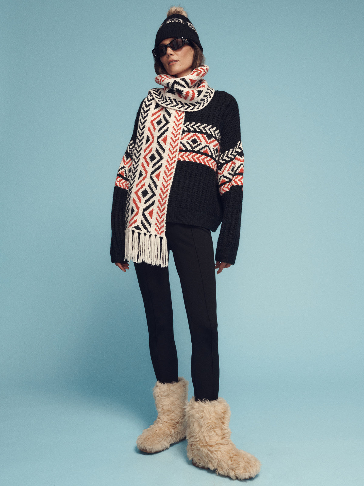 A person wearing the Sanctuary Clothing black multi intarsia scarf, a black and white patterned sweater, black pants, and large furry boots is set against a light blue background. They accessorize with sunglasses and a beanie, creating a cozy winter vibe.