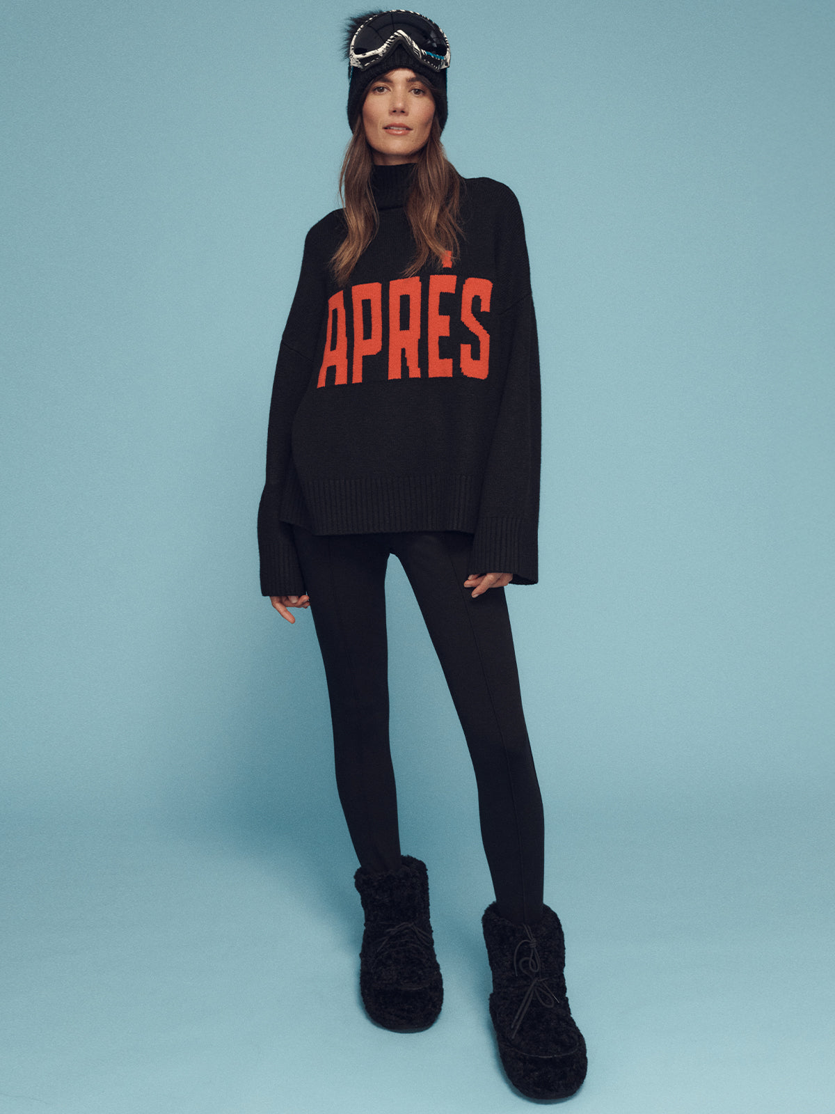 A person stands against a blue background wearing Sanctuary Clothing's Apres Intarsia Logo Sweater in black and mars red, paired with black leggings, furry black boots, a knit hat, and ski goggles resting on their head. Long hair cascades over their shoulders.