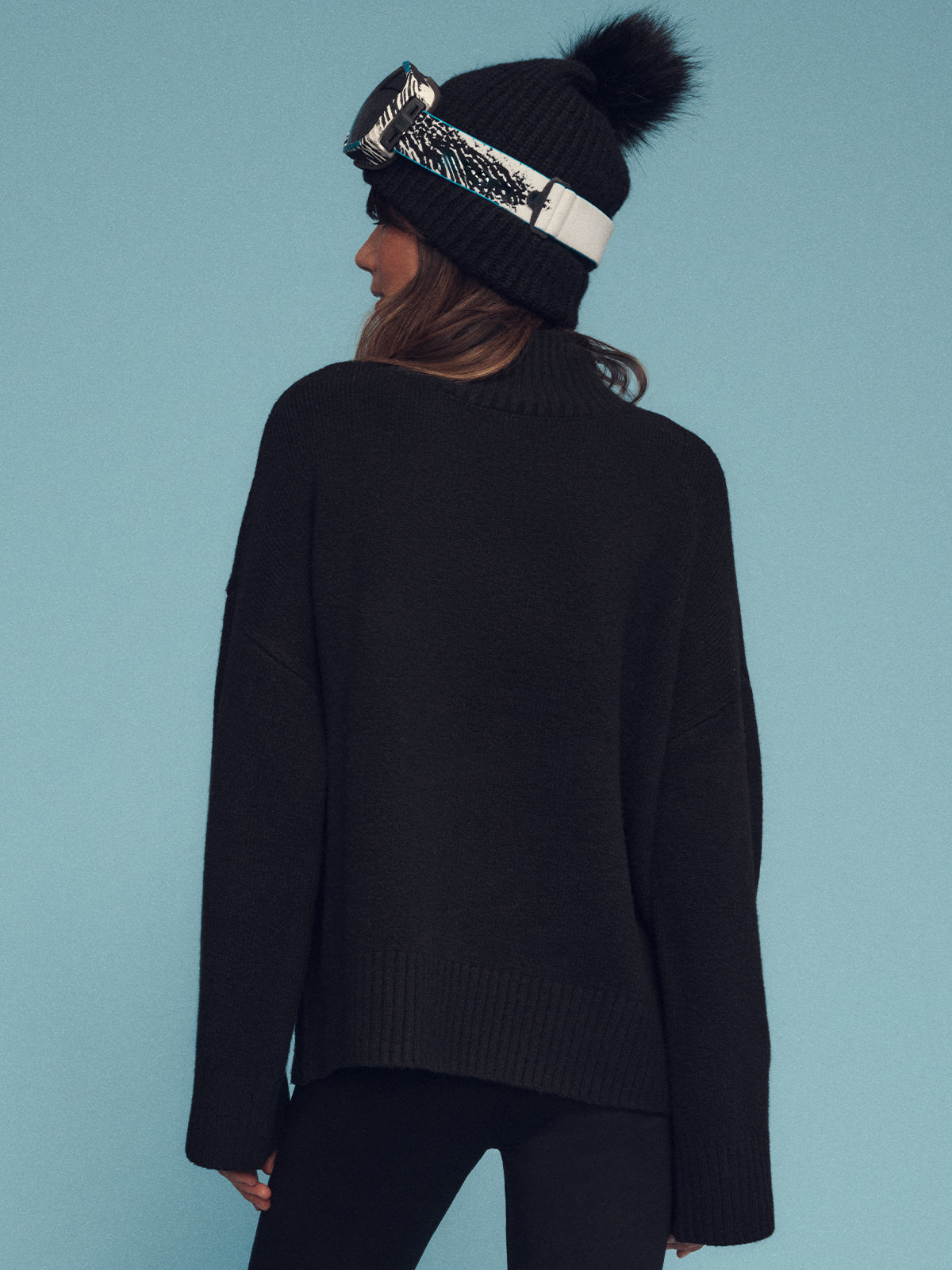 A person stands with their back to the camera, wearing a cozy "apres intarsia logo sweater black mars red" by Sanctuary Clothing, complemented by black leggings and a knitted black beanie adorned with a pompom. Ski goggles rest on the beanie, set against a plain blue background.