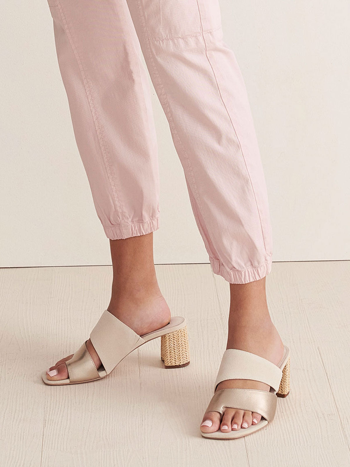 A person is standing on a light wooden floor, wearing light pink cuffed pants and the Brisk Sandal in Nude Bronze Sand Dollar by Sanctuary Clothing. The open-toe heels, featuring wide straps and a chunky, textured design, draw attention to the lower legs and feet.