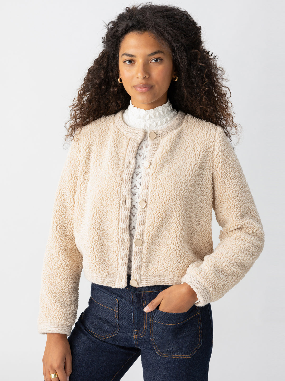 A person with long, curly hair is wearing Sanctuary Clothing's Cozy Cardigan in Toasted Almond over a white lace top. They have one hand in the pocket of their dark blue jeans and stand against a plain, light gray background.