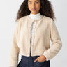 A person with long, curly hair is wearing Sanctuary Clothing's Cozy Cardigan in Toasted Almond over a white lace top. They have one hand in the pocket of their dark blue jeans and stand against a plain, light gray background.