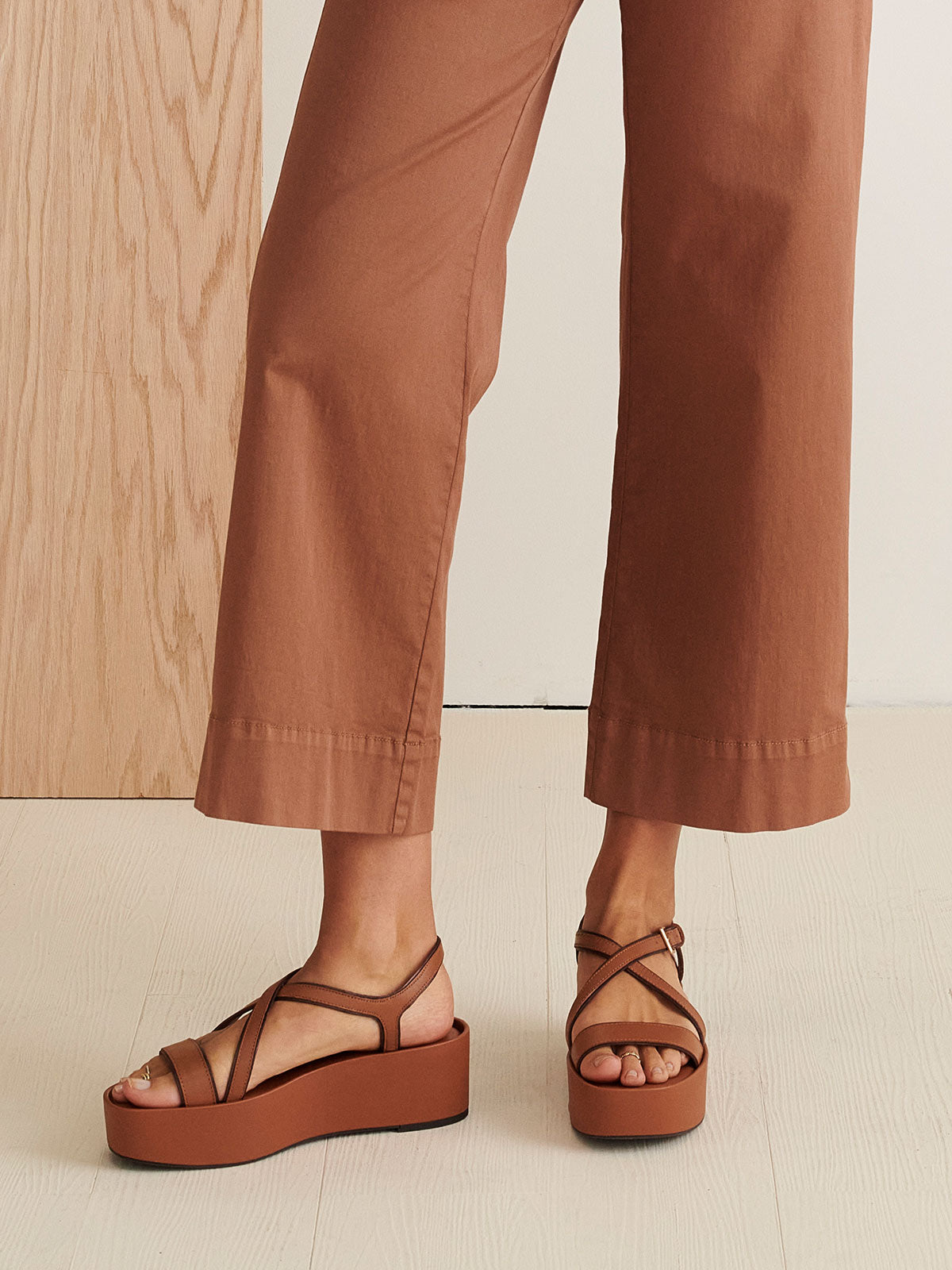 A person wearing brown wide-leg pants and the Define Sandal Deep Honey by Sanctuary Clothing stands on a light-colored wooden floor. A portion of a wood-paneled surface is visible in the background.