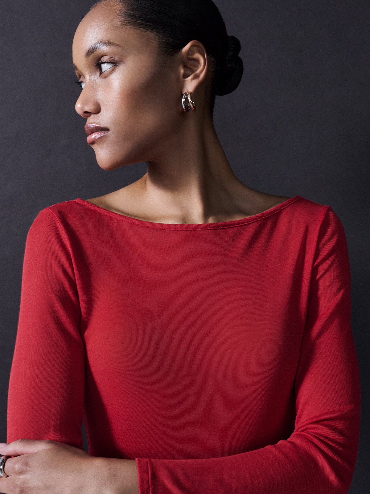 A person stands against a dark background, wearing a Sanctuary Clothing Semi Sheer Tee in Cherry Red and silver hoop earrings. They are looking to the left with an expression of calm contemplation, their hair neatly pulled back in a bun.