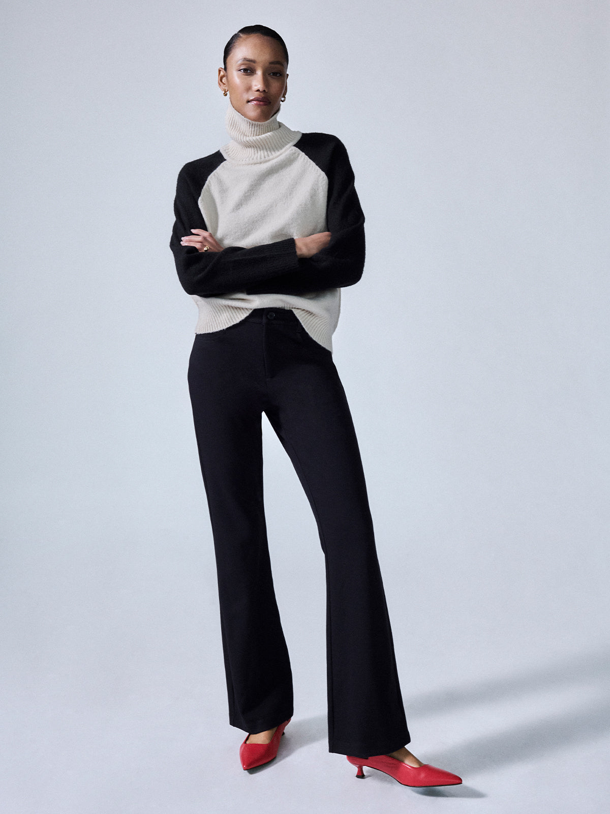 A person stands confidently with arms crossed, wearing the Maya Flare Black turtleneck sweater by Sanctuary Clothing, black pants, and red heeled shoes. The background is plain and minimalist.