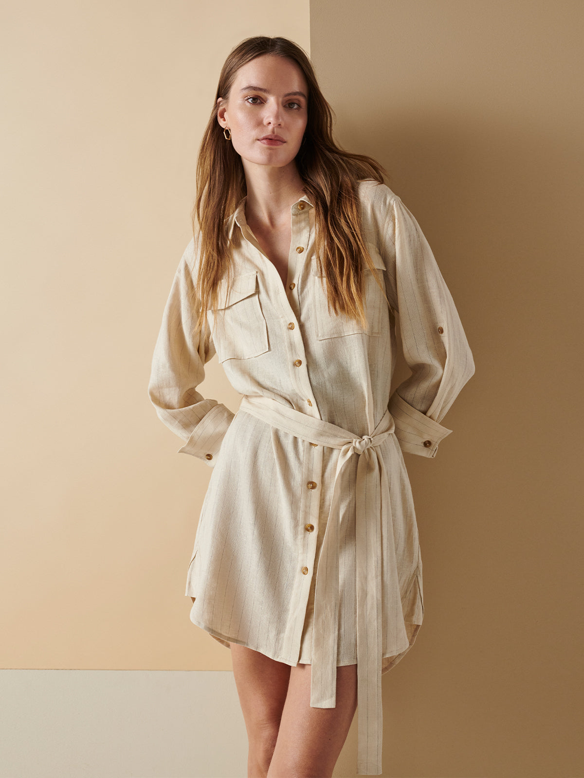 A woman with long hair stands against a beige wall, wearing the Pocket Shirt Dress Vineyard Stripe from Sanctuary Clothing, which features a light beige design, button-up front, waist tie, long sleeves and two front pockets. She is looking at the camera with a neutral expression.