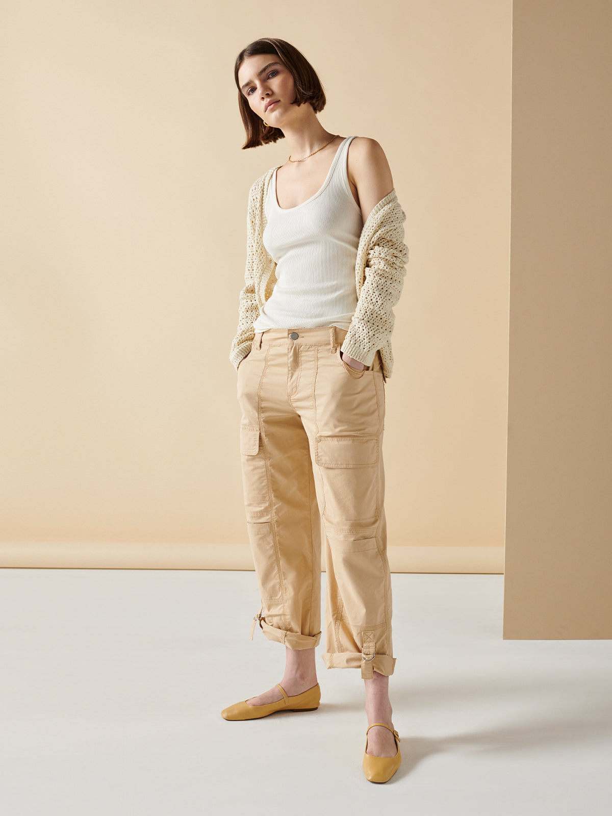 A person with short hair stands against a beige background, wearing the "My Kind Of Tank Birch" by Sanctuary Clothing, paired with a light knitted cardigan, beige cargo pants with multiple pockets, and yellow ballet flats. Their hands are in their pockets, and they have a relaxed pose.
