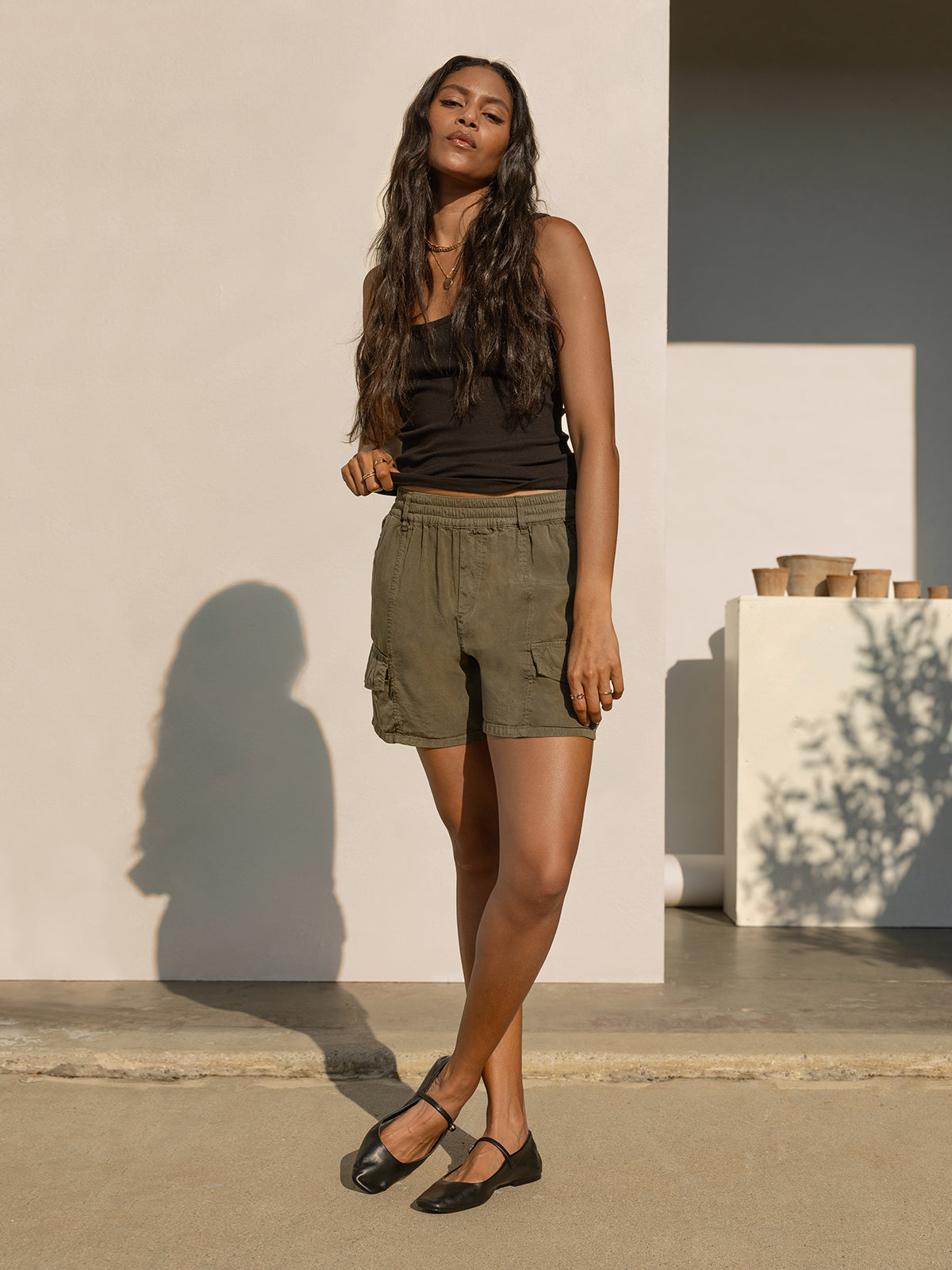 A woman with long wavy hair stands outside against a light-colored wall, wearing a black sleeveless top, the Relaxed Rebel Short in Burnt Olive from Sanctuary Clothing, and black shoes. Several pots are stacked on a white ledge in the background, with tree shadows cast on the wall.