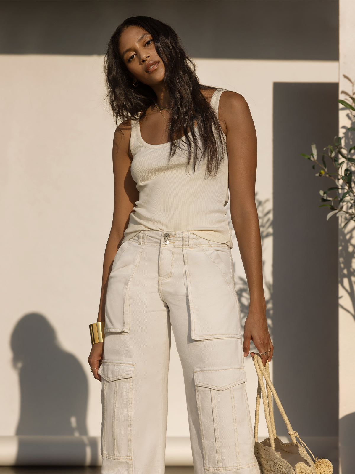 A woman stands against a plain wall, wearing a sleeveless beige top and the "Level Up Cargo Pant Powdered Sugar" from Sanctuary Clothing. She has long, wavy hair and holds a woven bag in one hand. Her look is completed with a gold bracelet, and she appears relaxed in the sunlight.