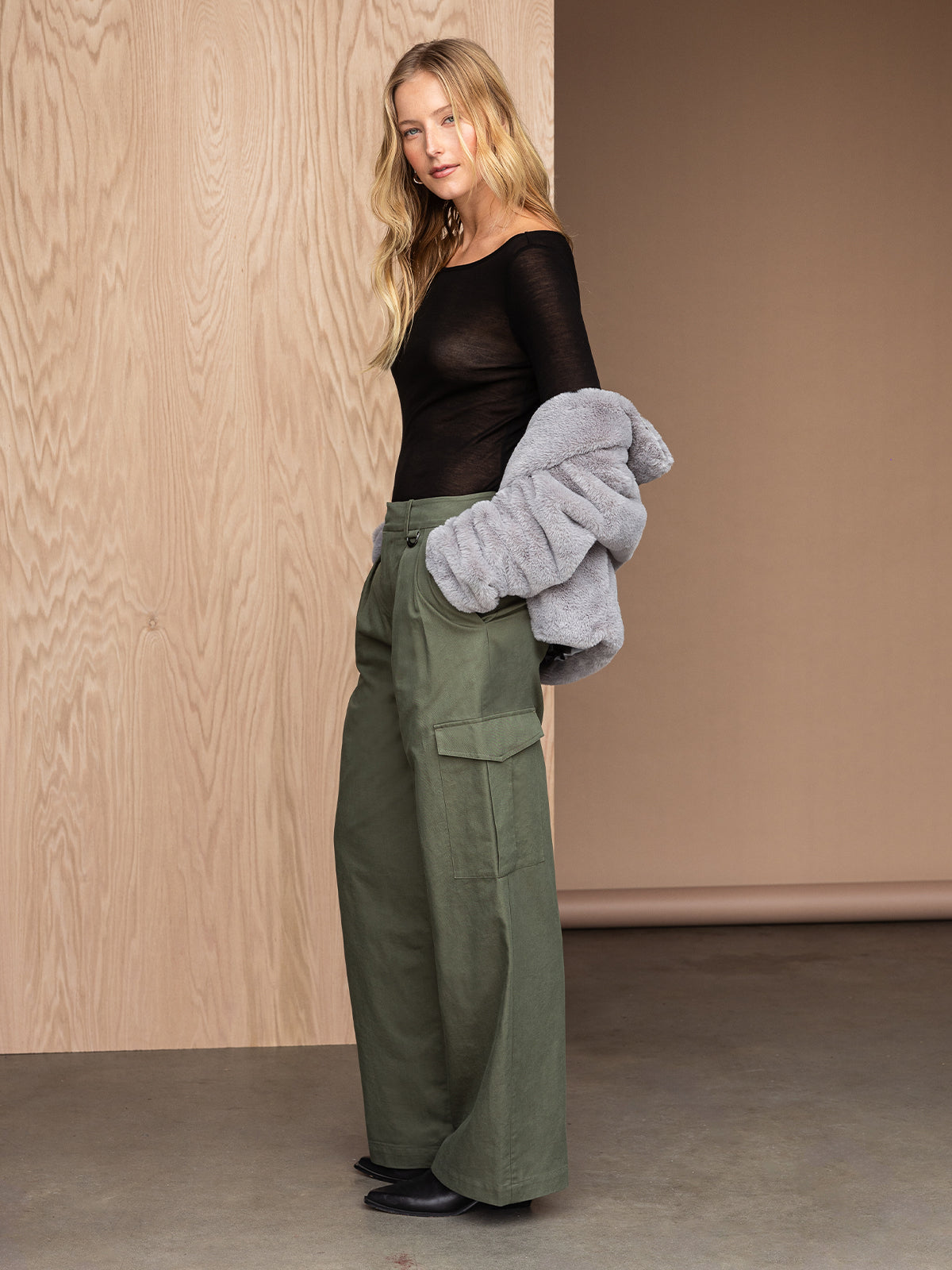 A woman with long blonde hair stands against a light wood-paneled wall. She wears a black sheer top, high-waisted Frankie Cargo Dark Olive pants by Sanctuary Clothing, and black shoes. She holds a gray jacket draped over her left arm and looks towards the camera with a neutral expression.