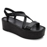 The Define Sandal Black Black by Sanctuary Clothing is a black wedge sandal featuring a wide toe strap and a crisscross ankle strap with a buckle. It has a thick sole with a textured pattern, offering both minimalist design and comfort.