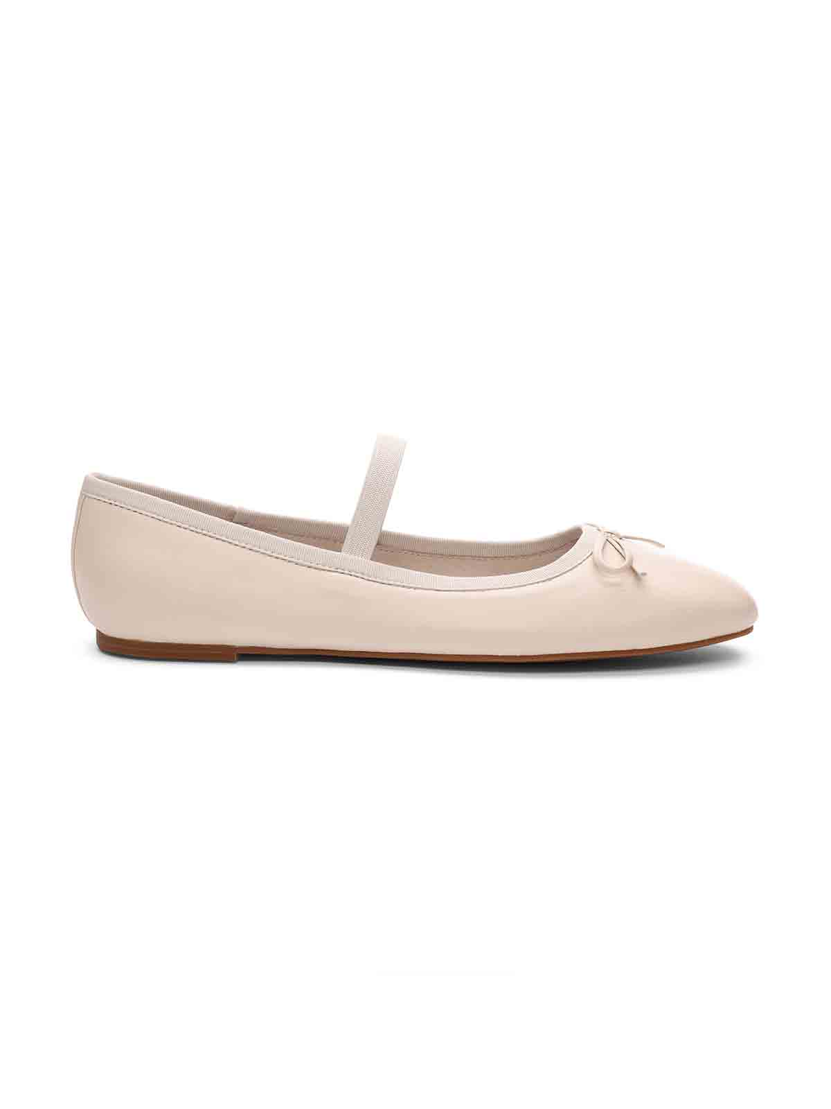 A side view of the "Facile Ballet Flat Milk" by Sanctuary Clothing, a beige ballet flat shoe featuring a thin elastic strap across the top. The shoe is adorned with a small bow on the front and has a rounded toe and flat heel. The material has a smooth and shiny finish.