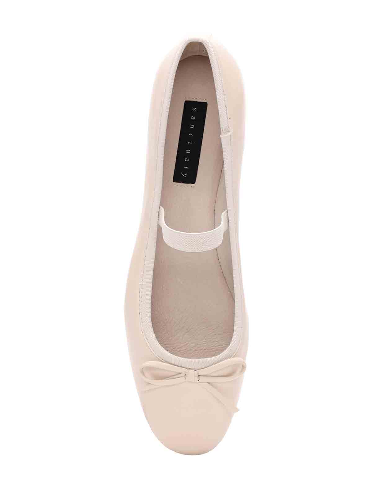 The Facile Ballet Flat Milk by Sanctuary Clothing features a beige ballet flat design with a small decorative bow on the toe and an elastic strap across the middle. The insole is branded "sanctuary." The shoe is positioned top-down against a white background.