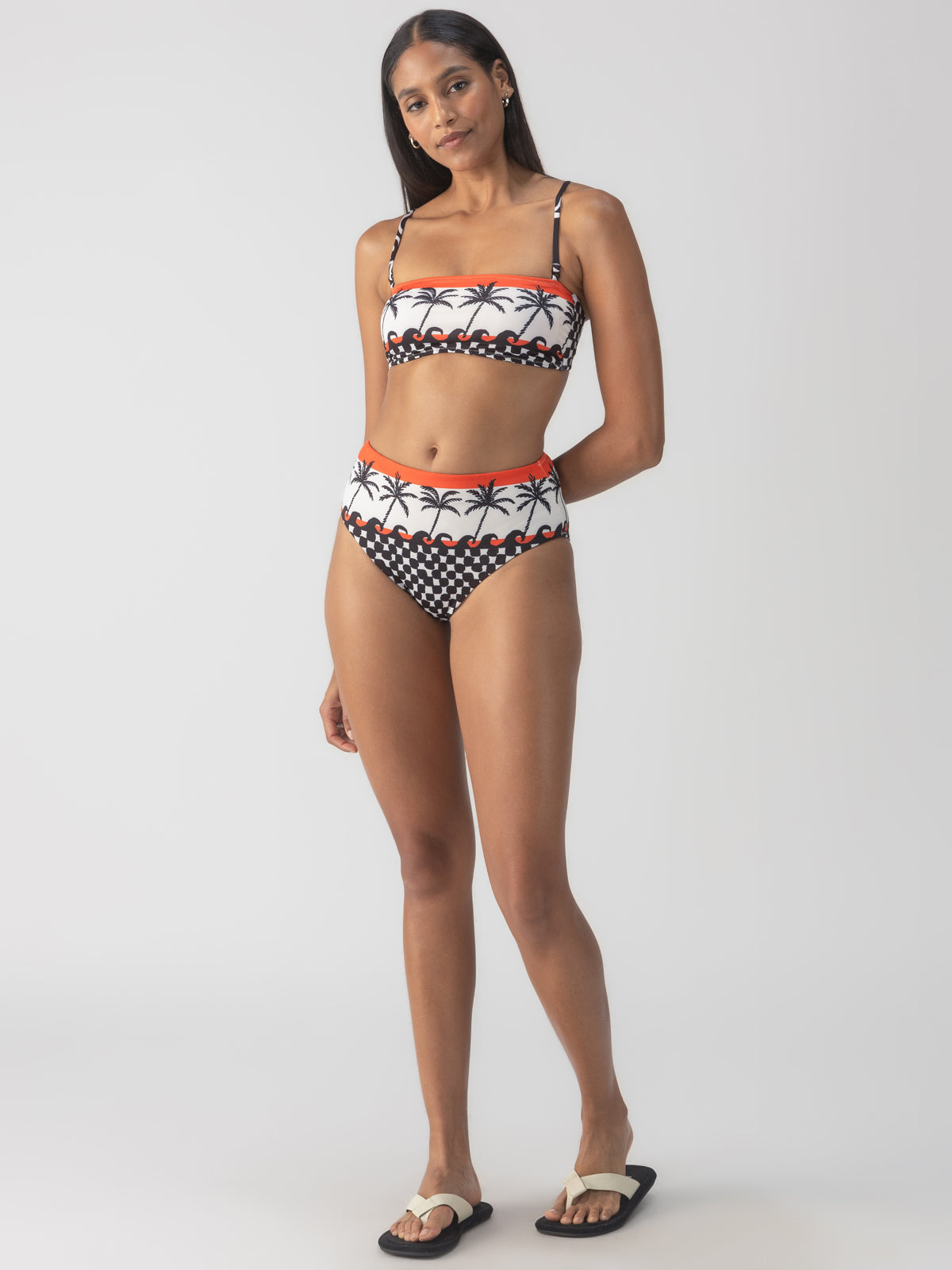 A person poses in a palm tree-patterned bikini with high-waisted bottoms, wearing the Sanctuary Clothing beach con reversible bandeau bikini top in black. They stand on a neutral background wearing black and white sandals.