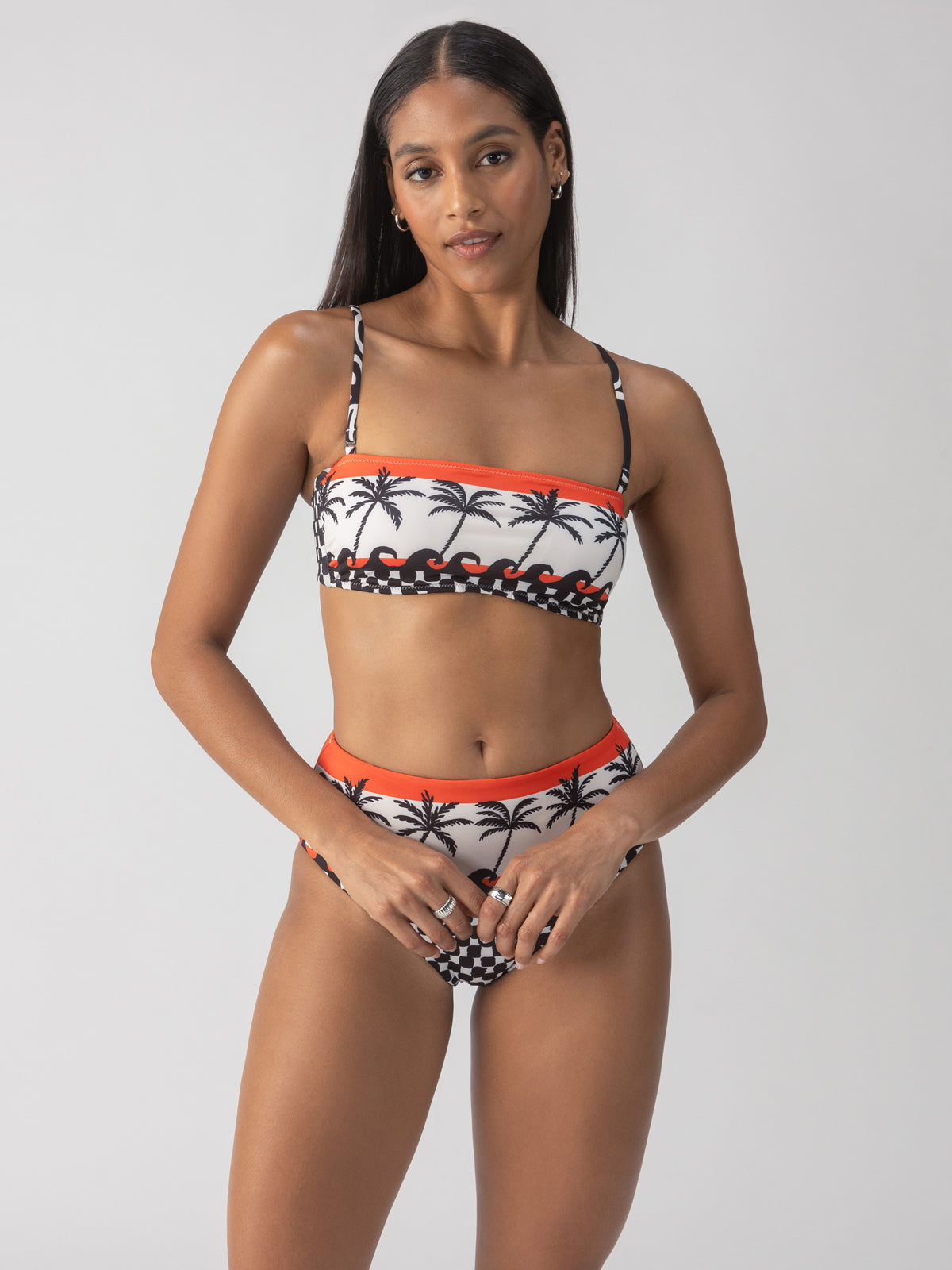 A person poses confidently wearing the "beach con reversible bandeau bikini top" by Sanctuary Clothing, adorned with palm tree and wave designs. The strapless top is paired with high-waisted bottoms highlighted with orange accents. The background is plain and light-colored.