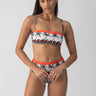 A person poses confidently wearing the "beach con reversible bandeau bikini top" by Sanctuary Clothing, adorned with palm tree and wave designs. The strapless top is paired with high-waisted bottoms highlighted with orange accents. The background is plain and light-colored.