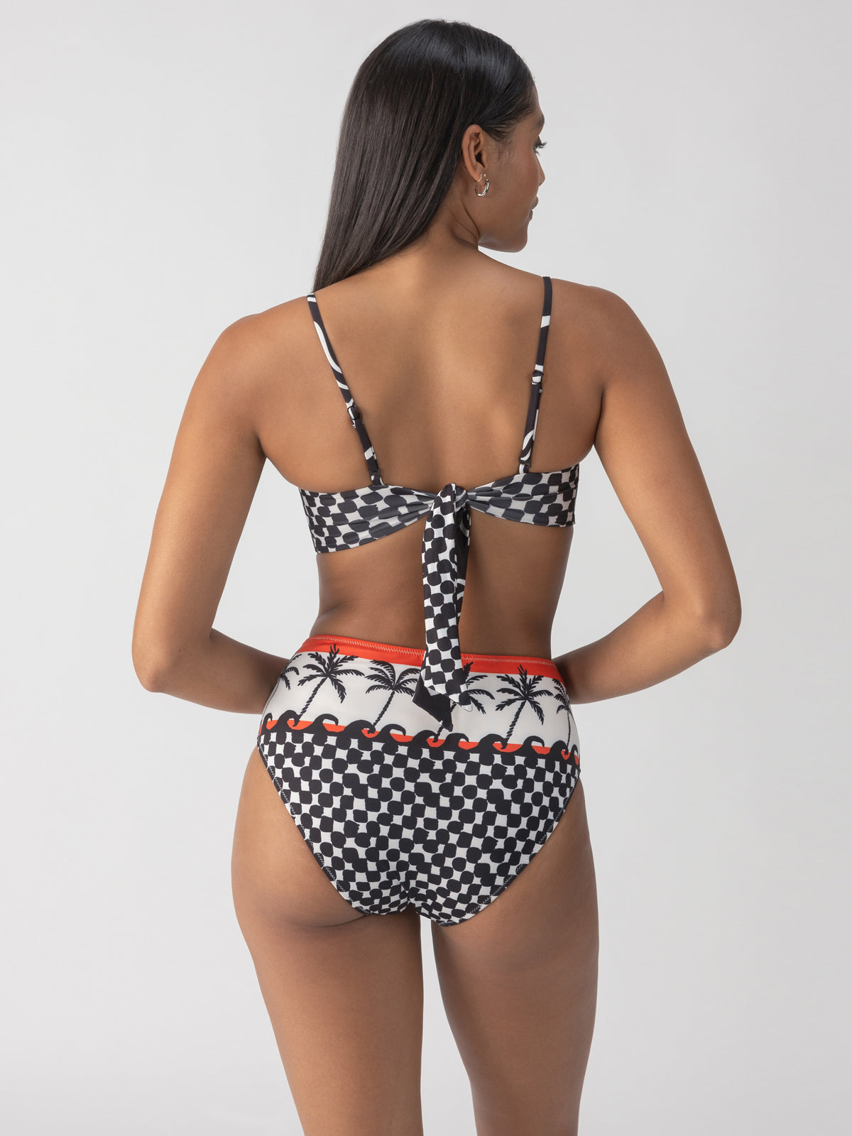 A woman with long black hair models the Sanctuary Clothing beach con reversible bandeau bikini top in black and white checkered design with palm trees and red accents. The image captures her from the back, showcasing the tie closure of the bikini top and the high-waisted style of the bottoms.