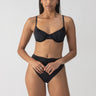 A person with long hair is standing confidently against a plain background, wearing the "Beach Goddess Excess Shirr Cup Bikini Top Black" from Sanctuary Clothing, paired with high-waisted bottoms. They have a calm expression and their hands are relaxed by their sides.