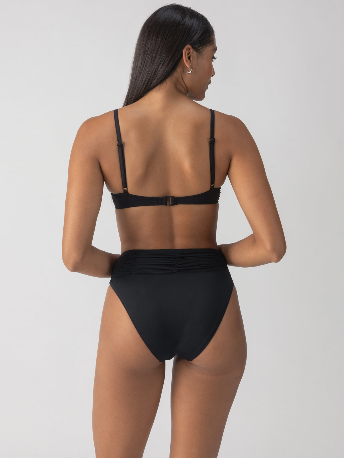 A woman is standing with her back to the camera, wearing a Sanctuary Clothing "beach goddess excess shirr cup" bikini top in black that features adjustable straps. The bikini has high-waisted bottoms, and the background is plain and light-colored.