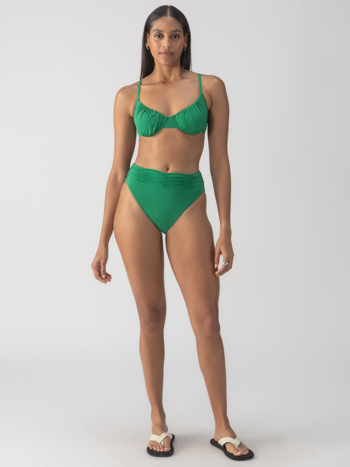 A person wearing the Beach Goddess Excess Shirr Cup Bikini Top in Perky Palm by Sanctuary Clothing stands against a plain background. They're wearing flip-flops and have long, straight hair.