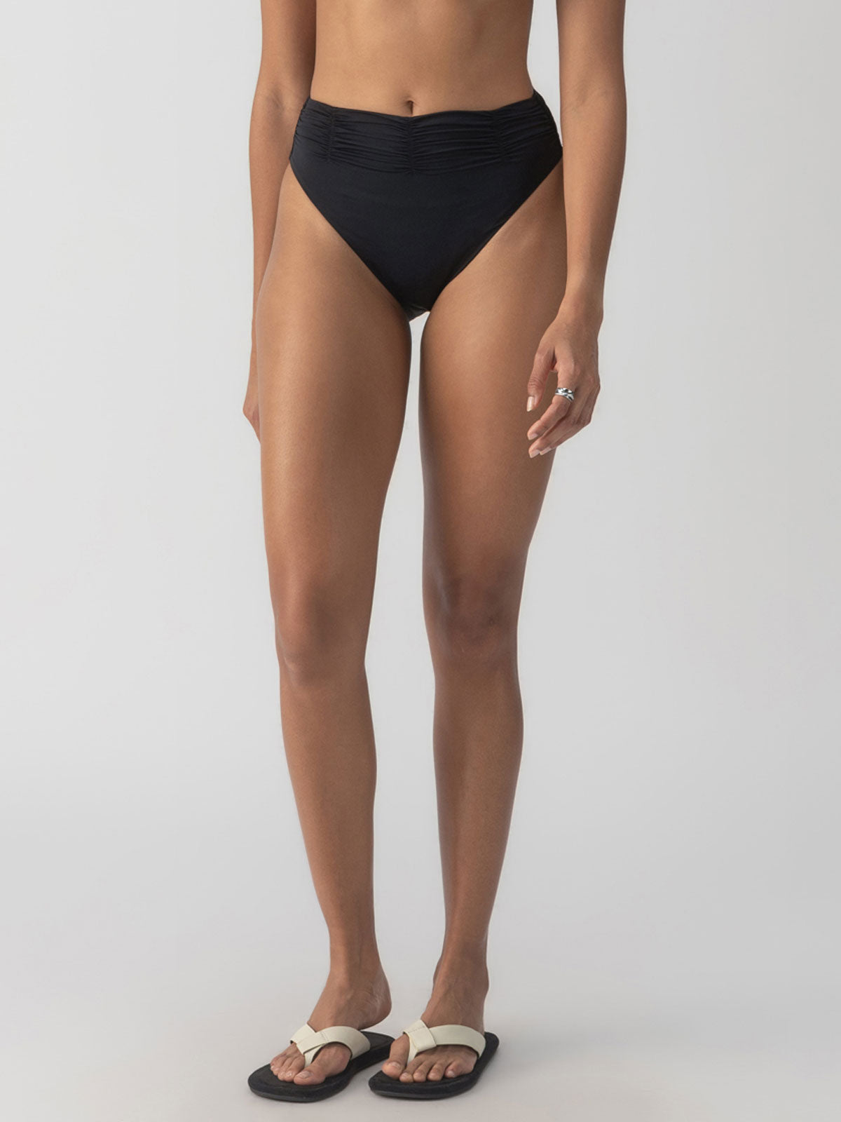 A person wearing the Beach Goddess Excess Shirr Waist Bikini Bottom in black from Sanctuary Clothing and sandals stands against a plain background, with a silver ring on their left hand.