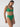 A person models the Sanctuary Clothing beach goddess excess shirr waist bikini bottom in "perky palm" against a plain background, looking downward. The green bikini features high-waisted bottoms and a ruched top with thin straps and gathered details.