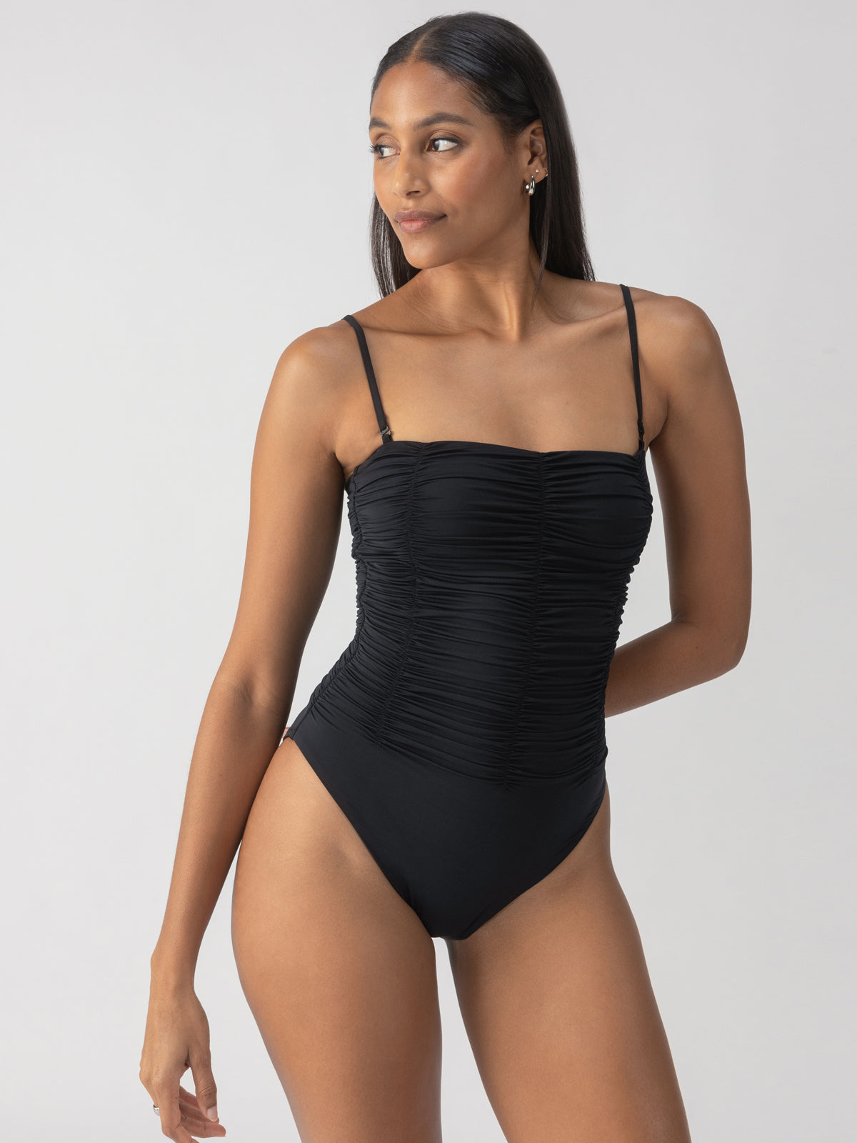 A person proudly poses against a neutral background, gazing to the side while wearing the Sanctuary Clothing Beach Goddess Excess Shirr Banded Mio One Piece in black, which highlights ruched detailing and adjustable straps.