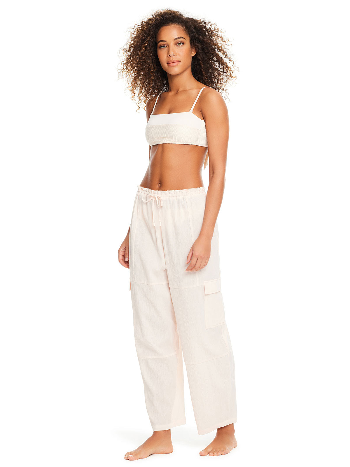 A woman with curly hair is standing and wearing the Sanctuary Clothing Coastal Cover Baggy Barrel Cargo Cover Up in White Sand. The light-colored, loose-fitting, sleeveless top and wide-legged pants with cargo pockets complement her relaxed style. She is barefoot, smiling slightly, and posing against a plain white background.