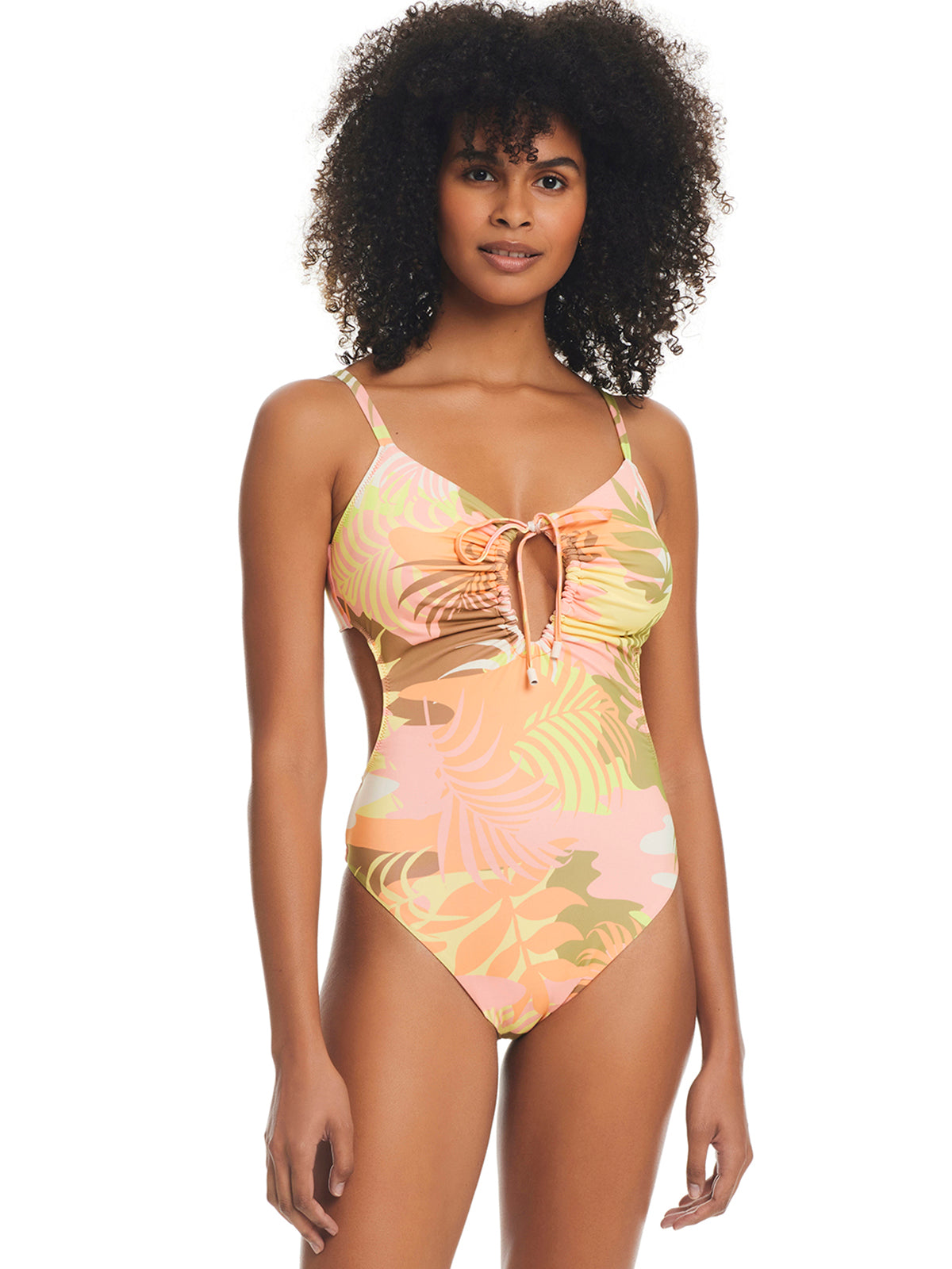 A woman with curly hair is wearing the Sanctuary Clothing Camo Palms Scoop Neck Monokini Multi, a colorful one-piece swimsuit featuring a tropical leaf print. The swimsuit showcases a keyhole cutout in the front with a tie detail. She stands against a plain white background, facing forward with a relaxed expression.