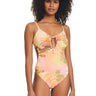 A woman with curly hair is wearing the Sanctuary Clothing Camo Palms Scoop Neck Monokini Multi, a colorful one-piece swimsuit featuring a tropical leaf print. The swimsuit showcases a keyhole cutout in the front with a tie detail. She stands against a plain white background, facing forward with a relaxed expression.