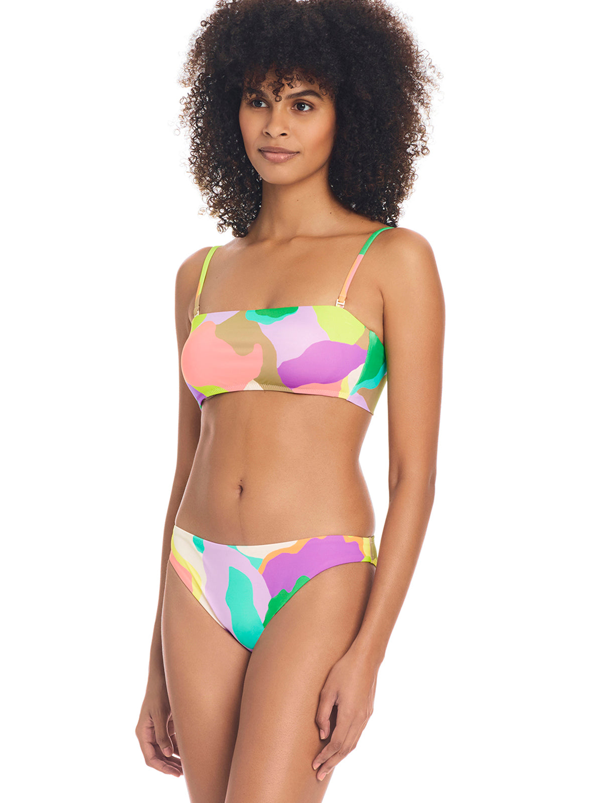 A woman with curly hair is wearing the Daytripper Bandeau Bikini Top Multi from Sanctuary Clothing, featuring a colorful, abstract pattern. She pairs it with matching bikini bottoms and stands while looking slightly off to the side, maintaining a neutral expression against a plain white background.