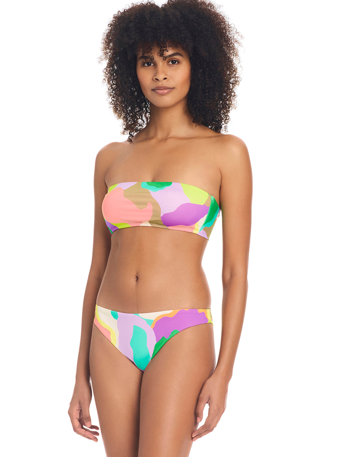 A person with curly hair is wearing a colorful, abstract-patterned bikini from Sanctuary Clothing. The swimwear includes the Daytripper Hipster Bikini Bottom Multi and features a bandeau top and low-rise bottoms. The background is plain white.