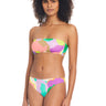 A person with curly hair is wearing a colorful, abstract-patterned bikini from Sanctuary Clothing. The swimwear includes the Daytripper Hipster Bikini Bottom Multi and features a bandeau top and low-rise bottoms. The background is plain white.