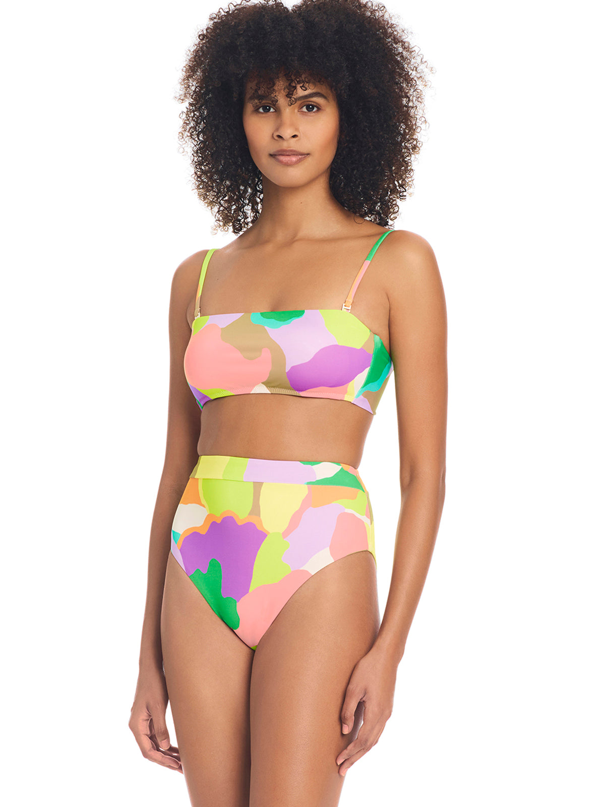 A person with curly hair is wearing a vibrant Daytripper High Rise Bikini Bottom Multi from Sanctuary Clothing, featuring a colorful camouflage pattern with hues of pink, yellow, green, and purple. The set includes a high-waisted bottom and a square-neck top against a white background.