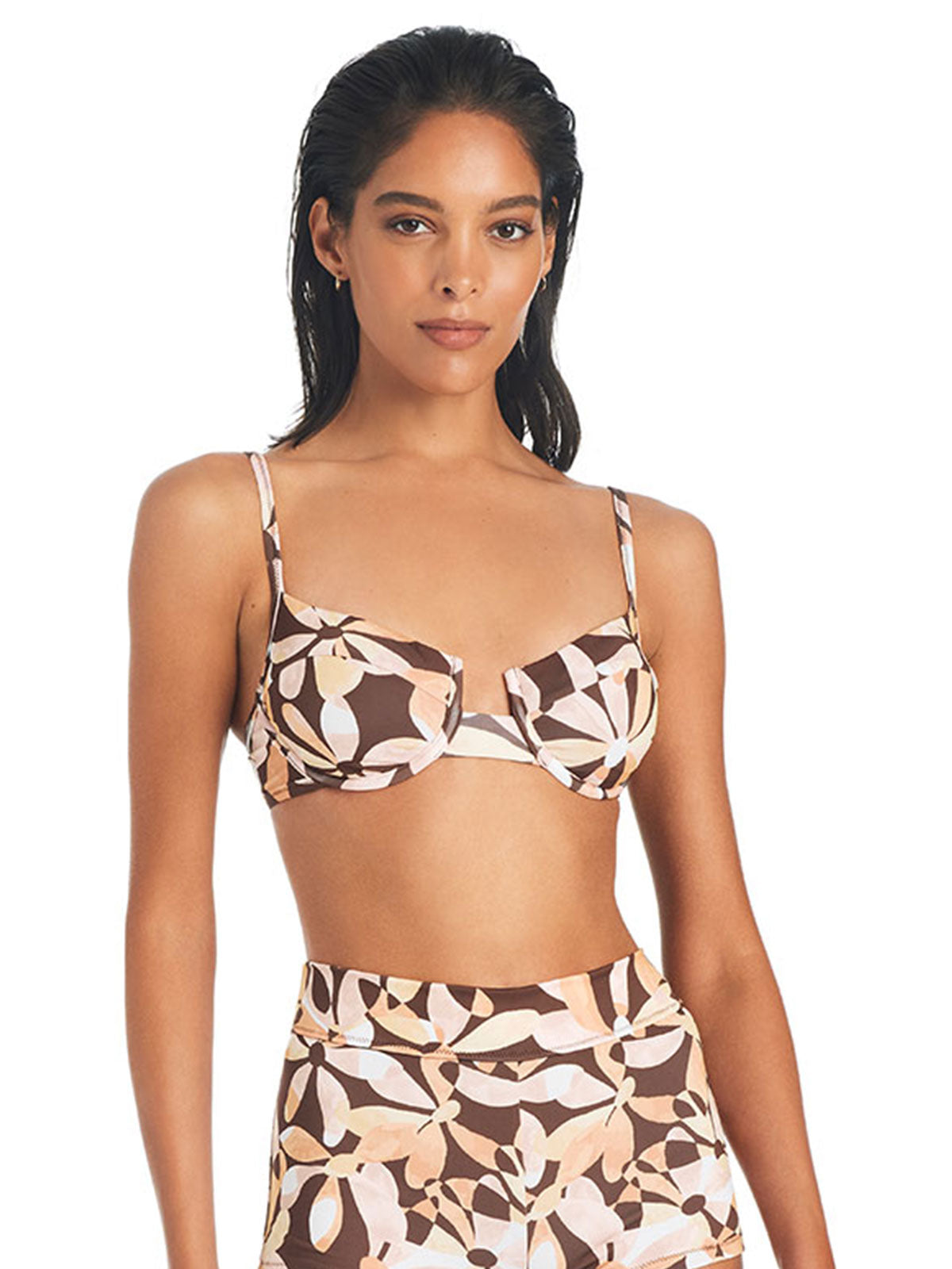 A woman with long dark hair wears the Jigsaw Petals Bikini Top Chocolate by Sanctuary Clothing, featuring a brown, peach, and white abstract floral pattern. The top has thin straps and underwire support, and she pairs it with high-waisted bottoms showcasing the same vibrant design. She stands against a plain white background.