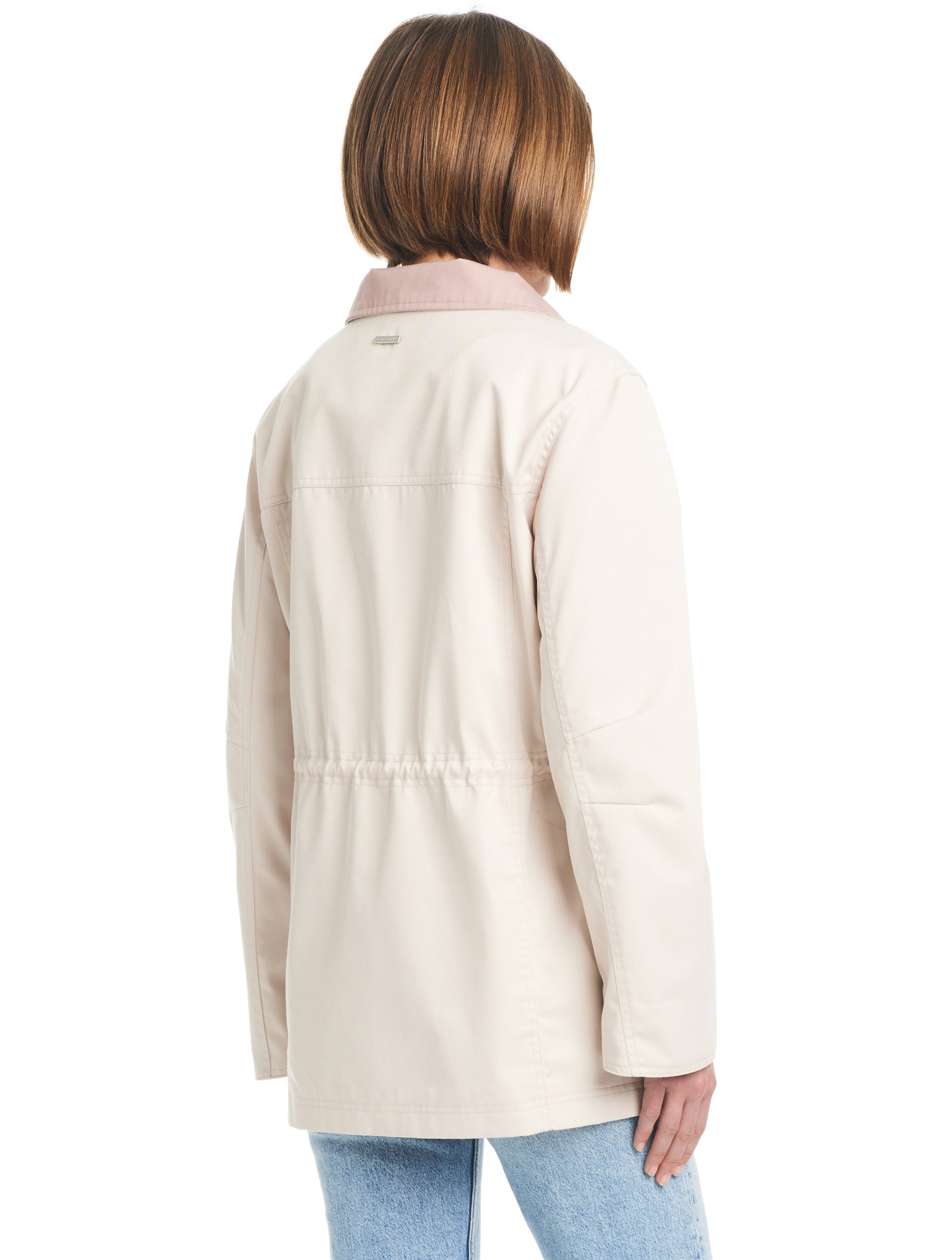 A person with short brown hair is standing with their back to the camera. They are wearing Sanctuary Clothing's Cotton Touch Relaxed Twill Jac in Moonbeam, a light beige jacket with a drawstring waist, paired with light blue jeans. The background is white.