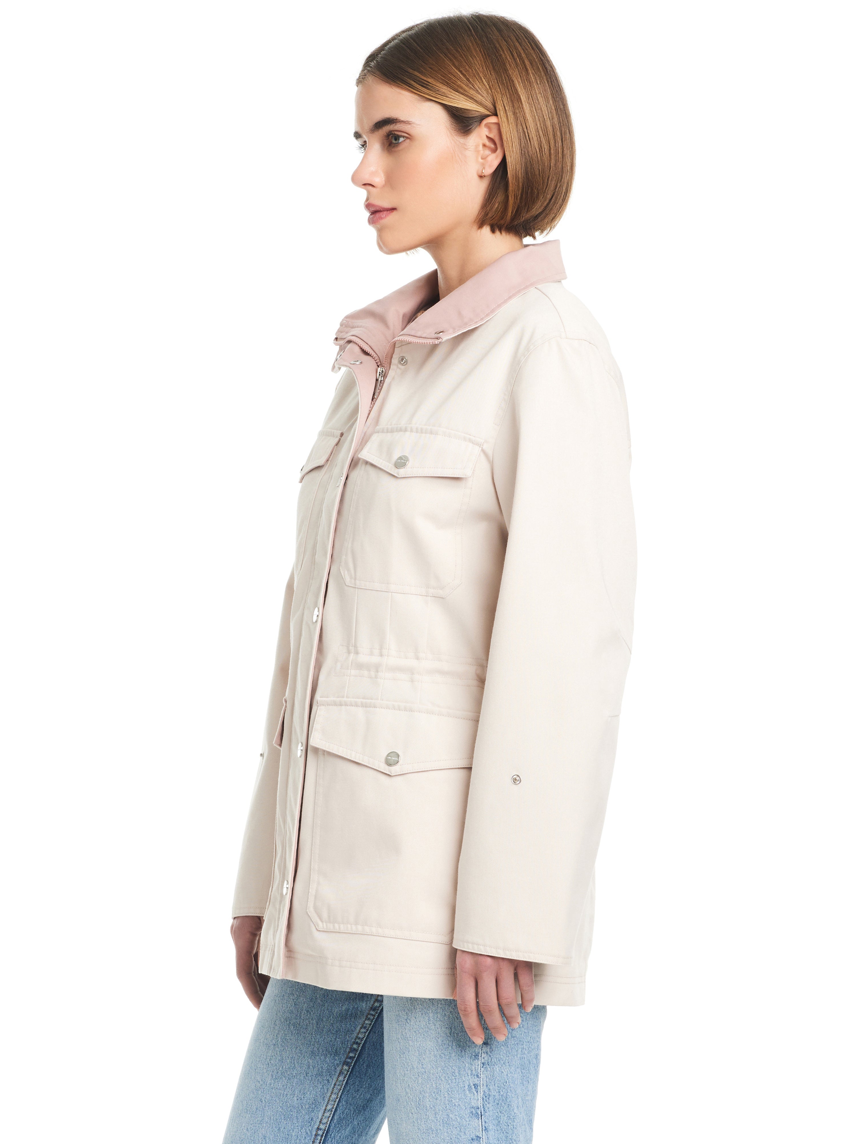 A woman with short, straight hair is standing in profile, wearing the Cotton Touch Relaxed Twill Jac Moonbeam jacket from Sanctuary Clothing. The light-colored jacket features large pockets and snap buttons on the front. She is also wearing light blue jeans against a plain white background.