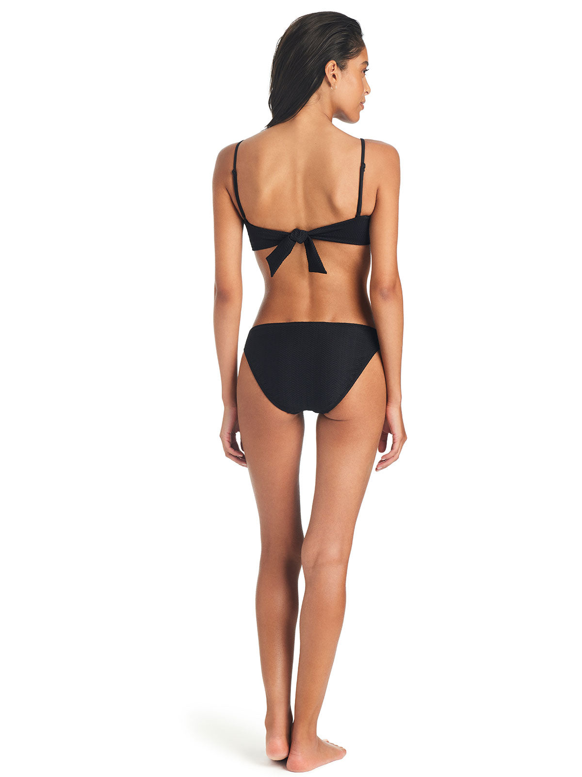 A woman with long dark hair stands with her back to the camera, showcasing the Sanctuary Clothing On The Water Texture Hipster Bikini Bottom in black. The bikini top features a bow at the back, and the bottoms are designed in a classic cut. She is posed against a plain white background.