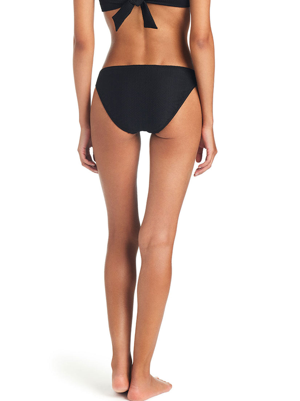 A woman is standing barefoot, facing away from the camera, wearing the Sanctuary Clothing On The Water Texture Hipster Bikini Bottom in black. The image highlights her back and legs, with the bikini top tied in a bow at the back. The background is plain white.