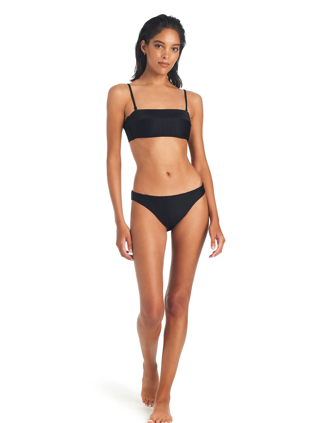 A woman is standing with one foot slightly forward, wearing the On The Water Texture Bandeau Bikini Top Black from Sanctuary Clothing, paired with mid-rise bottoms. The background is plain white, and she has long, dark hair. The overall look is minimalist and stylish.