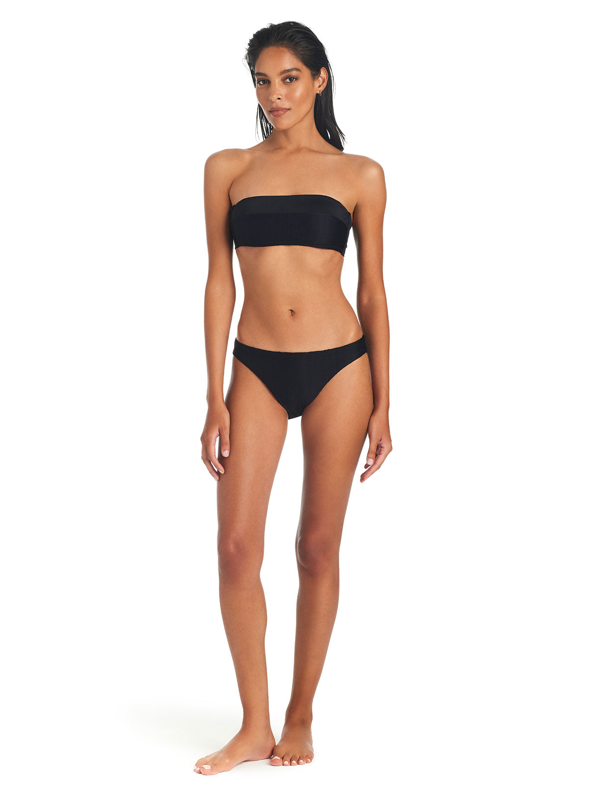 A woman stands barefoot against a white background, wearing the "On The Water Texture Bandeau Bikini Top Black" by Sanctuary Clothing paired with black bikini bottoms. She has long dark hair and a neutral expression, with her arms resting naturally by her sides.